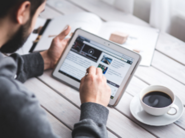 Mann mit Tablet in der Hand sitzt am Tisch mit einer Kaffeetasse neben sich